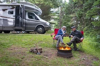 two adults sitting next to a fire next to an rv