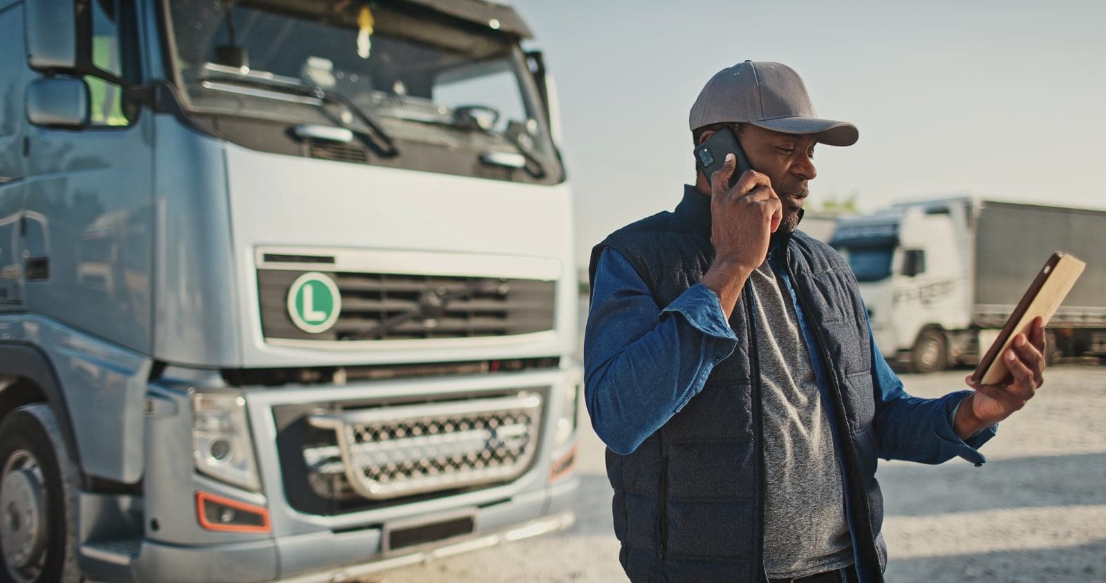 Truck driver on the phone with a tablet beside parked tractors, sorting out an Amazon Relay insurance certificate with RMIS